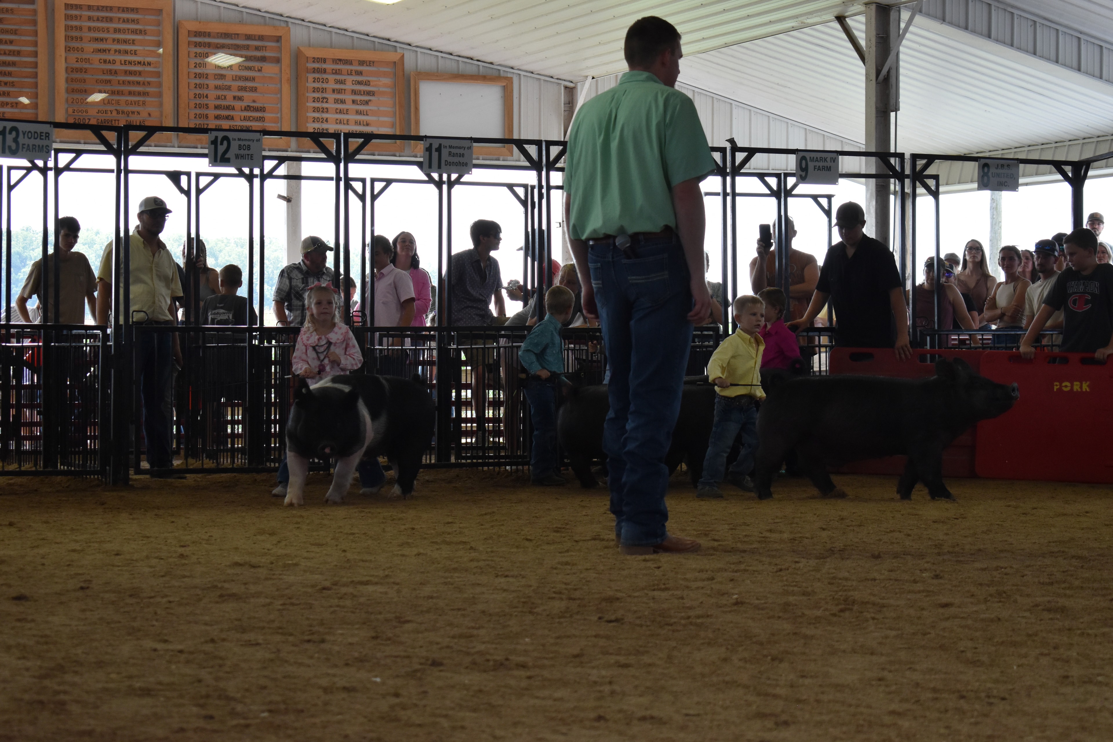Wide arena shot with judge in green shirt watching girl in pink floral top and boy in yellow shirt steer spotted Hampshire cross pig with packed sponsor pens 9–13 and past champions boards behind