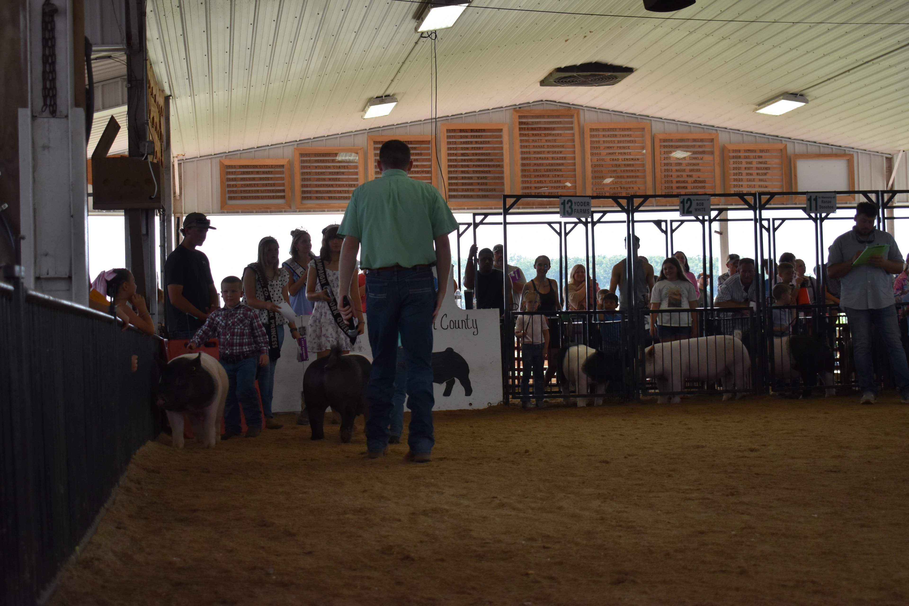Wide back-of-arena shot with judge in green shirt, Pork Queen with sash, and small boy in plaid shirt steering spotted Hampshire cross pig near the Champaign County Pork sign with packed crowd in Yoder Farms pens 11–13 and past champions boards behind