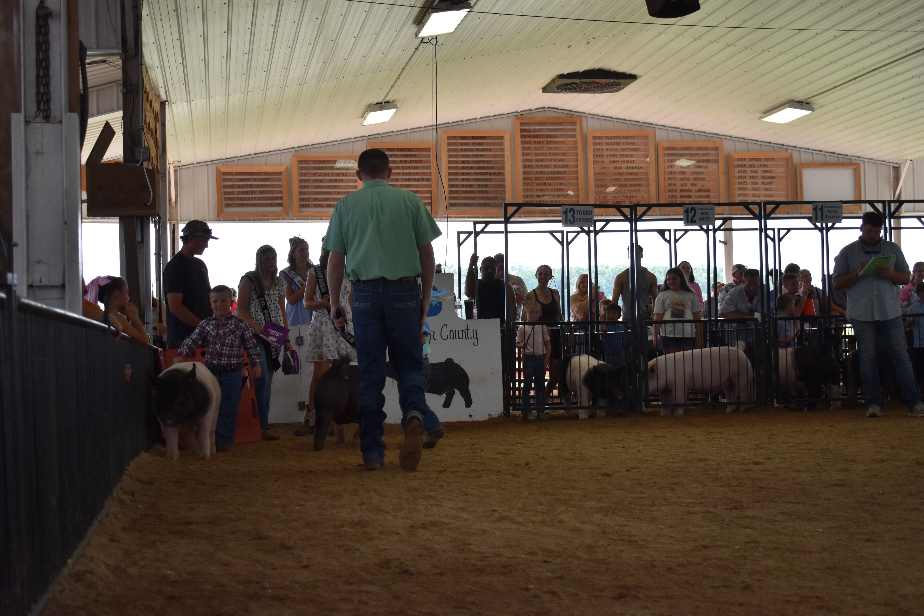 Wide back-of-arena shot with judge in green shirt bending toward small boy in plaid shirt steering spotted Hampshire cross pig near the Champaign County Pork sign with Pork Queen and packed crowd in Yoder Farms pens 11–13 behind