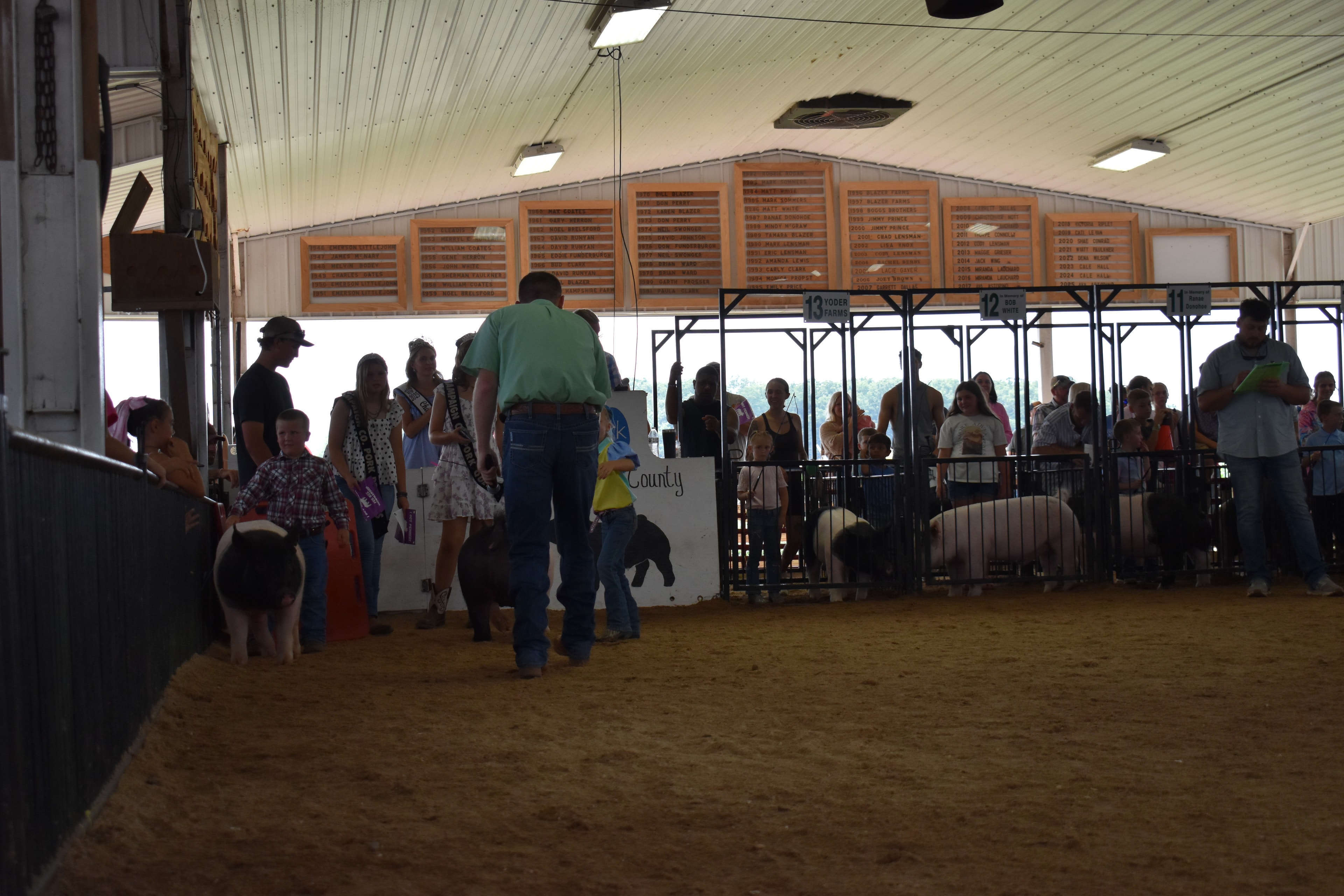 Wide back-of-arena shot with judge in green shirt, Pork Queen with sash, and small boy in plaid shirt steering spotted Hampshire cross pig near the Champaign County Pork sign with packed crowd in Yoder Farms pens 11–13 and past champions boards behind