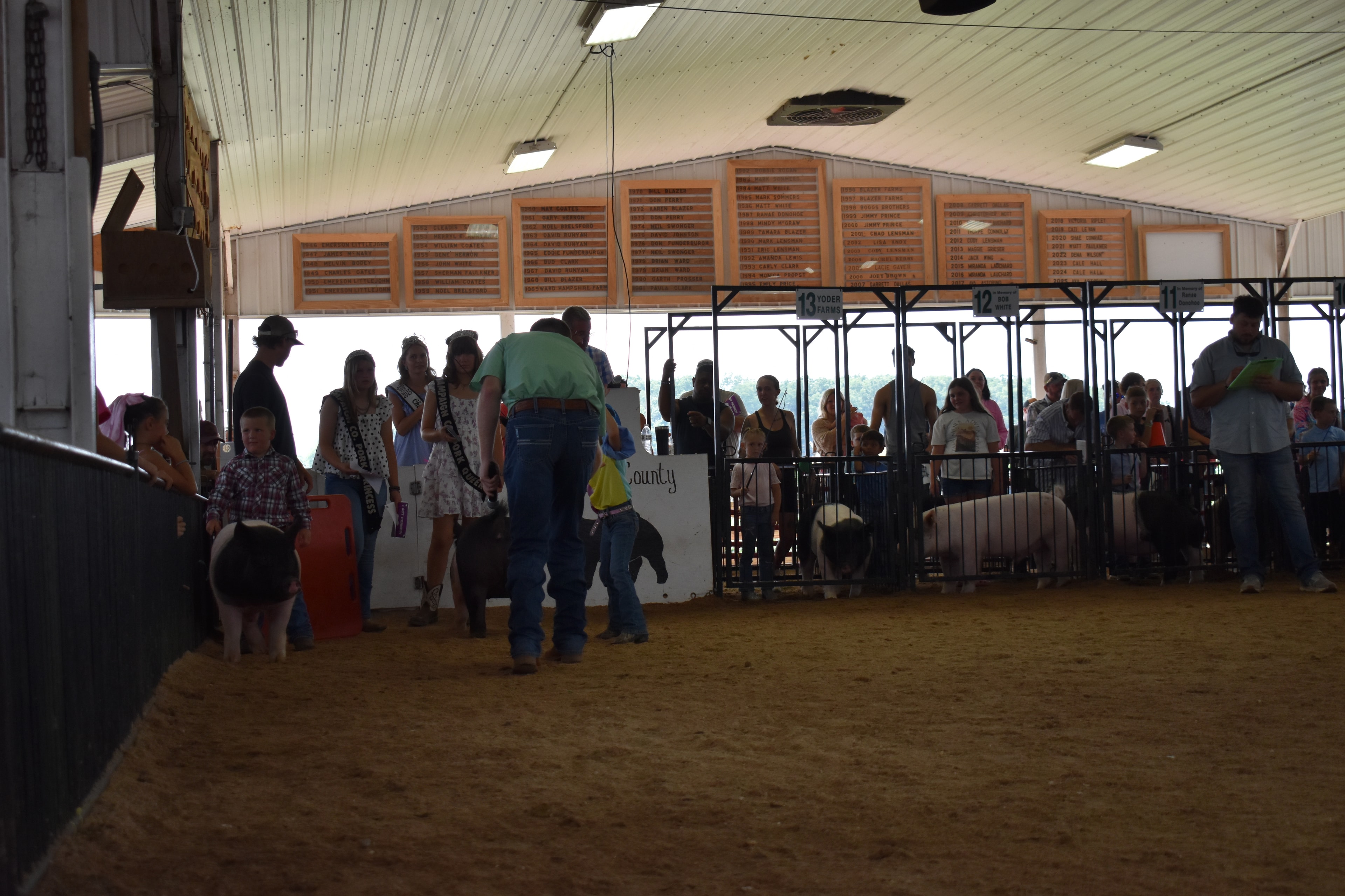 Wide back-of-arena shot with judge in green shirt, Pork Queen with sash, and small boy in plaid shirt steering spotted Hampshire cross pig near the Champaign County Pork sign with packed crowd in Yoder Farms pens 11–13 and past champions boards behind