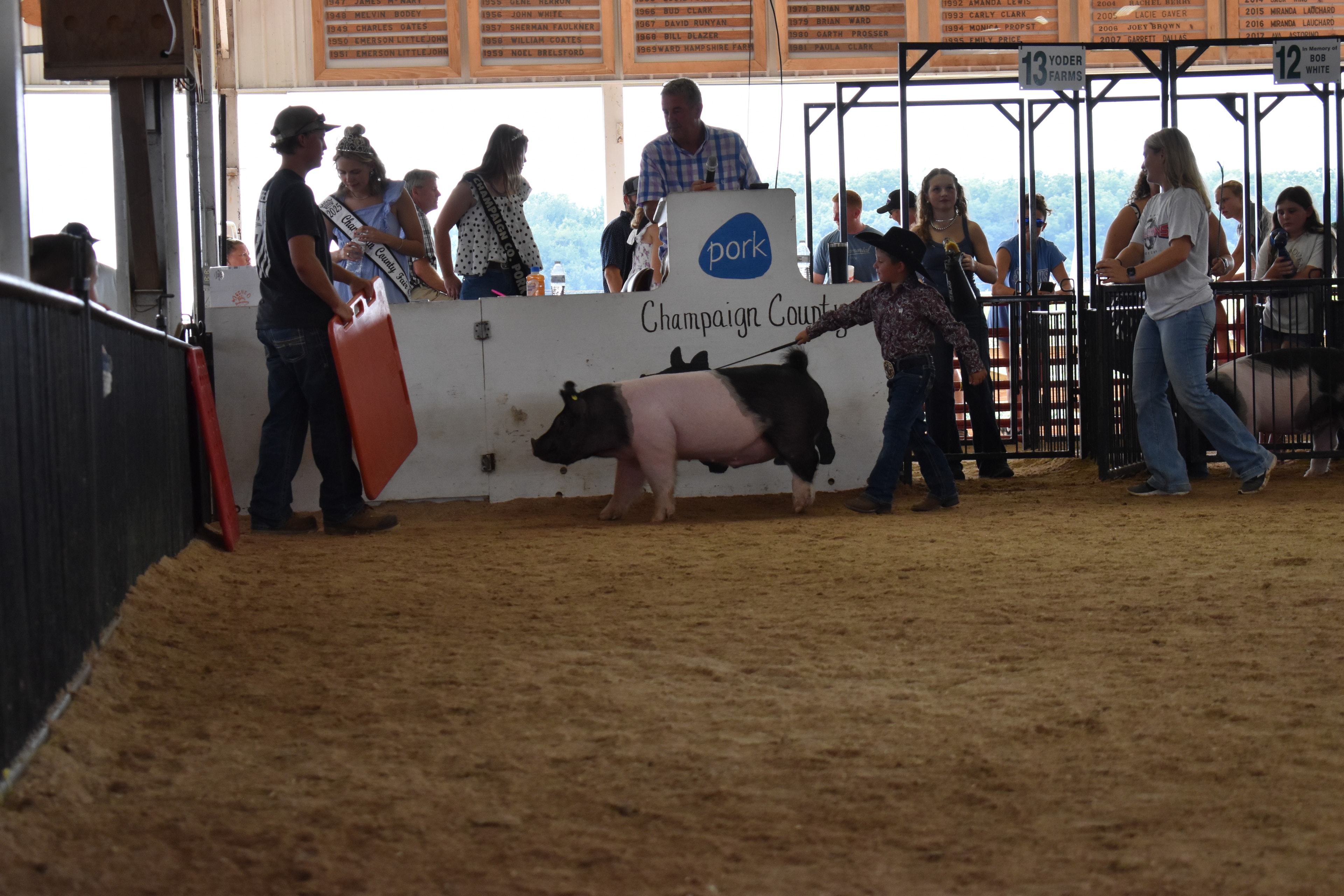 Small boy in black cowboy hat steering Hampshire cross pig sideways past Champaign County Pork sign with past champions boards