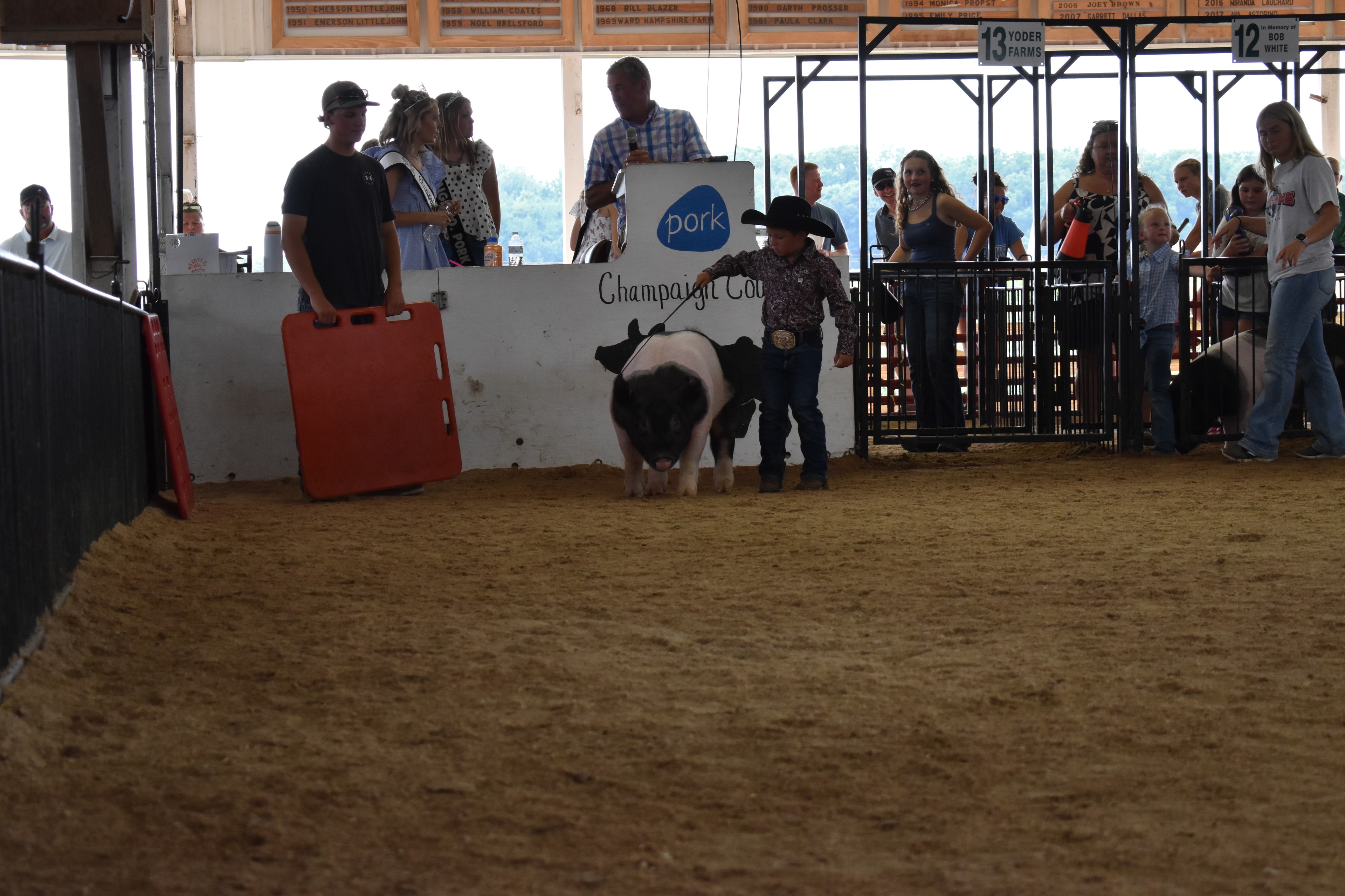 Small boy in black cowboy hat steering Hampshire cross pig past Champaign County Pork sign with Pork Queen and crowd