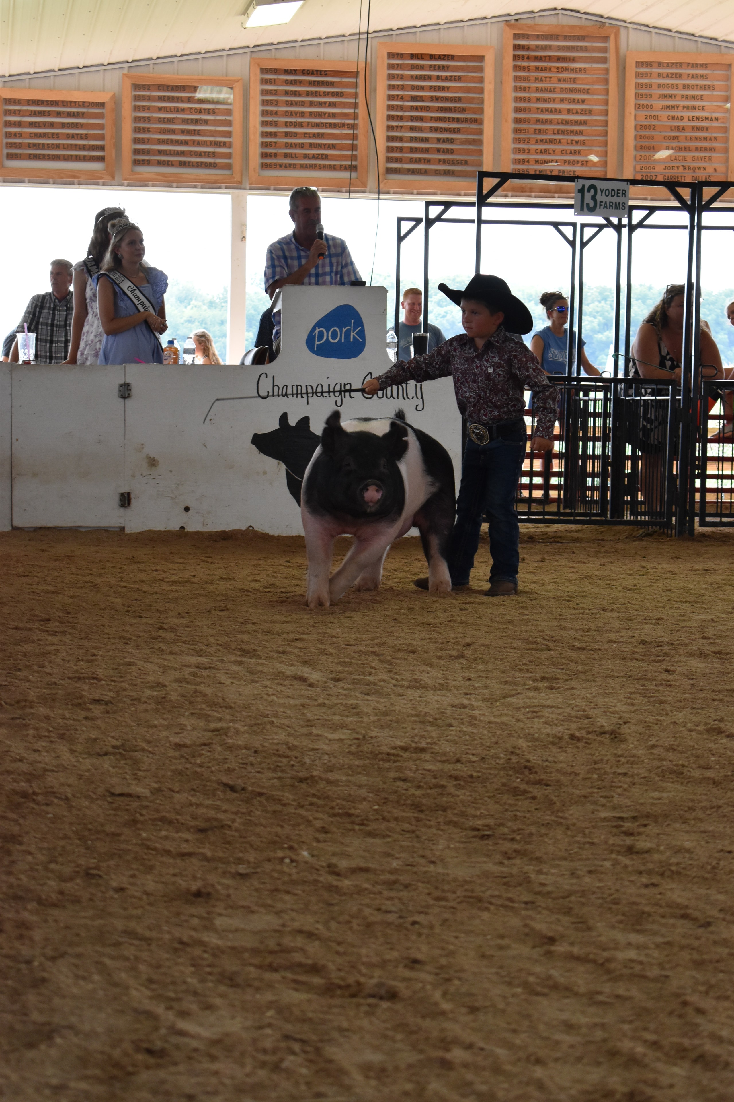 Small boy in black cowboy hat steering Hampshire cross pig past Champaign County Pork sign with Pork Queen and crowd watching