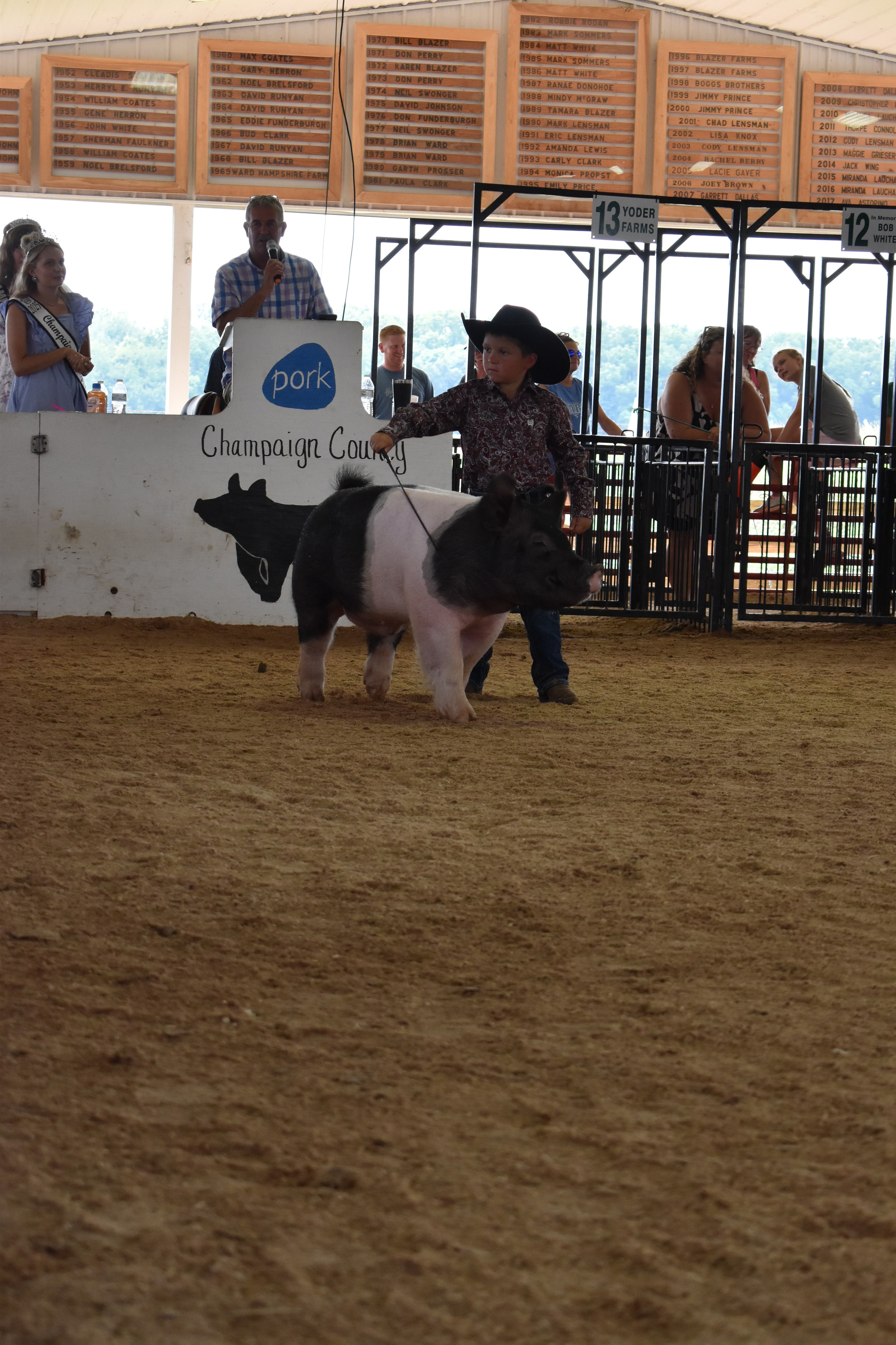 Boy in paisley shirt and black cowboy hat steering large Hampshire cross pig past Champaign County Pork sign with past champions boards