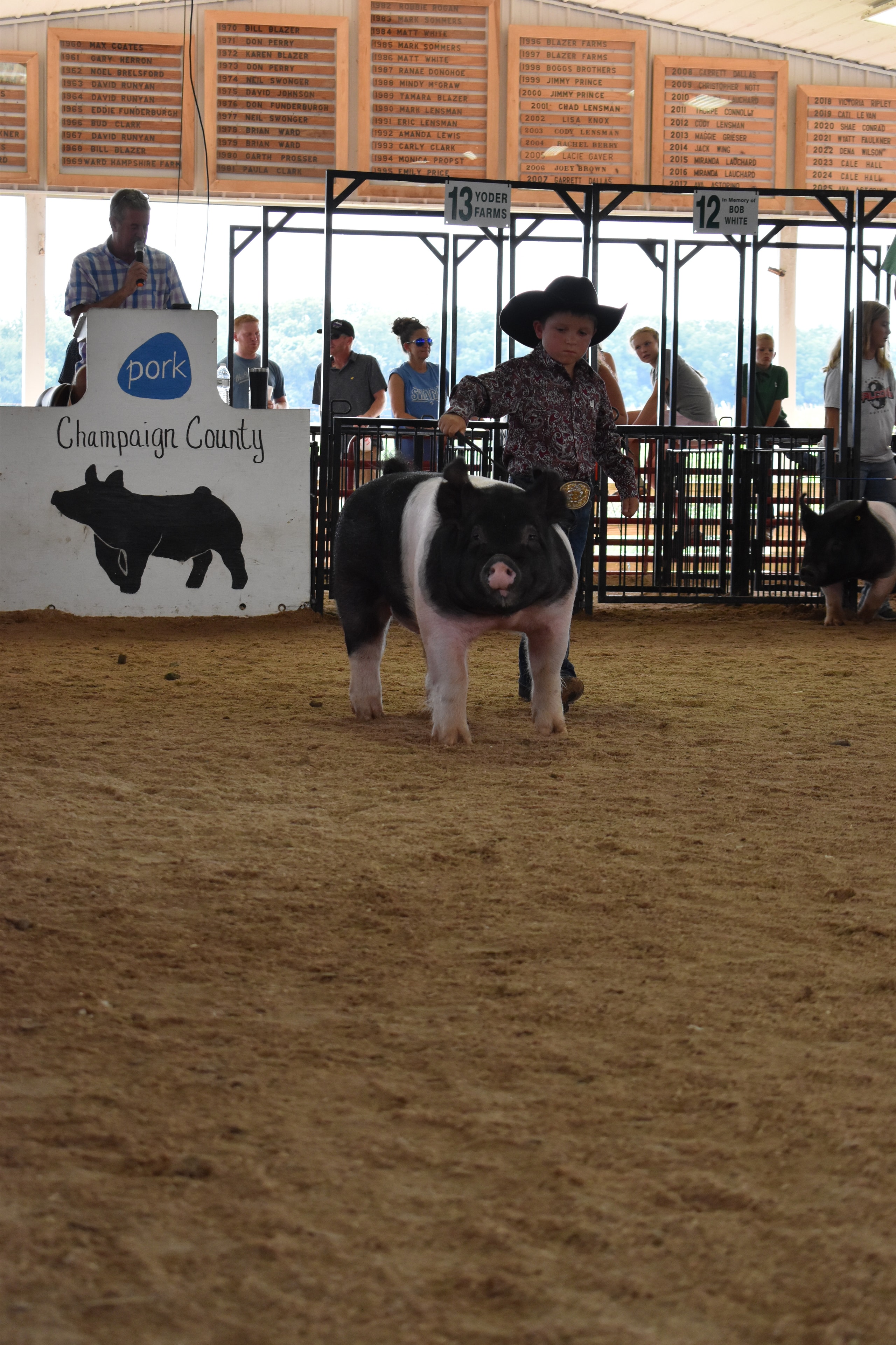 Boy in paisley shirt and black cowboy hat steering Hampshire cross pig facing camera with Champaign County Pork sign behind