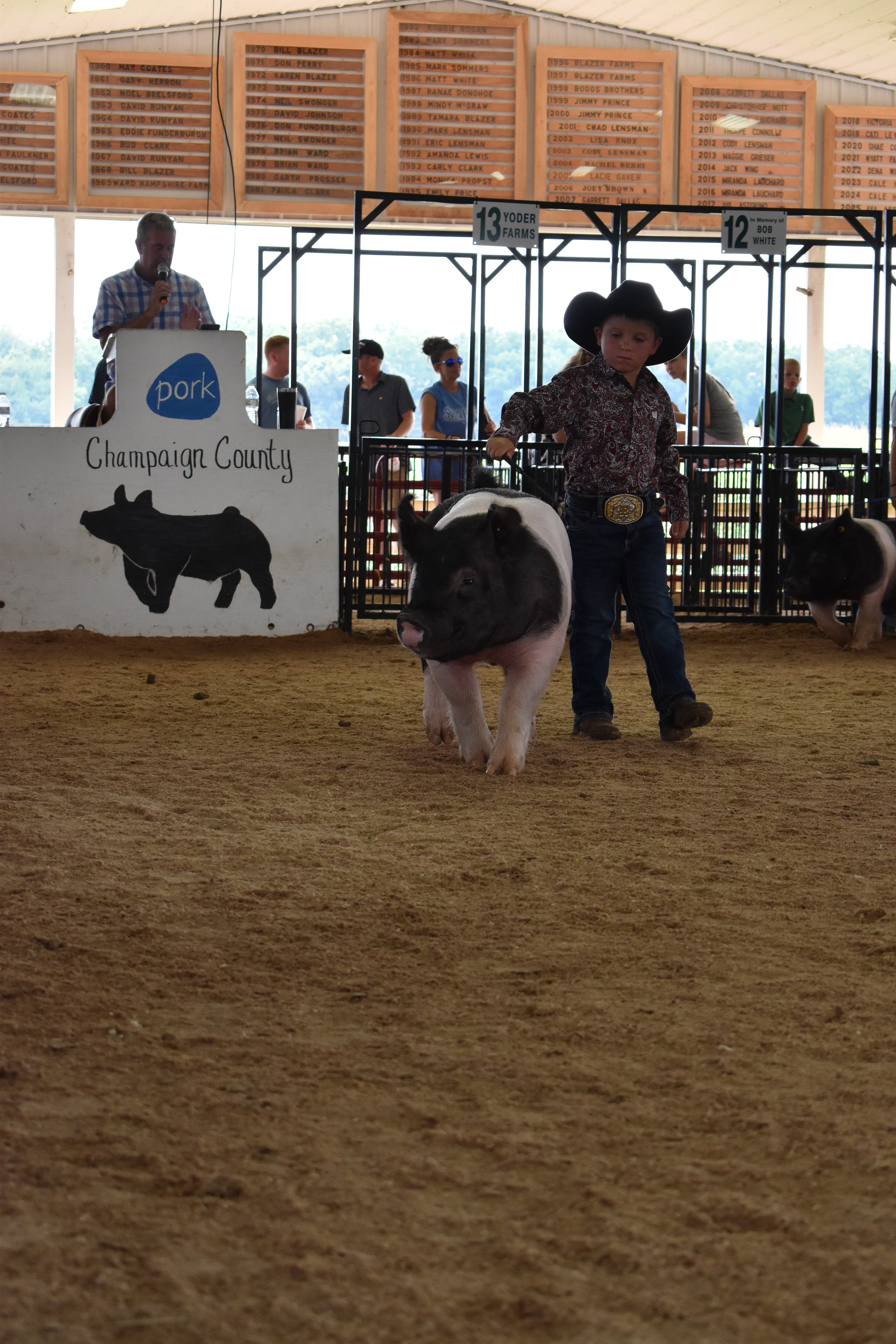 Boy in paisley shirt and black cowboy hat steering Hampshire cross pig sideways past Champaign County Pork sign with past champions boards
