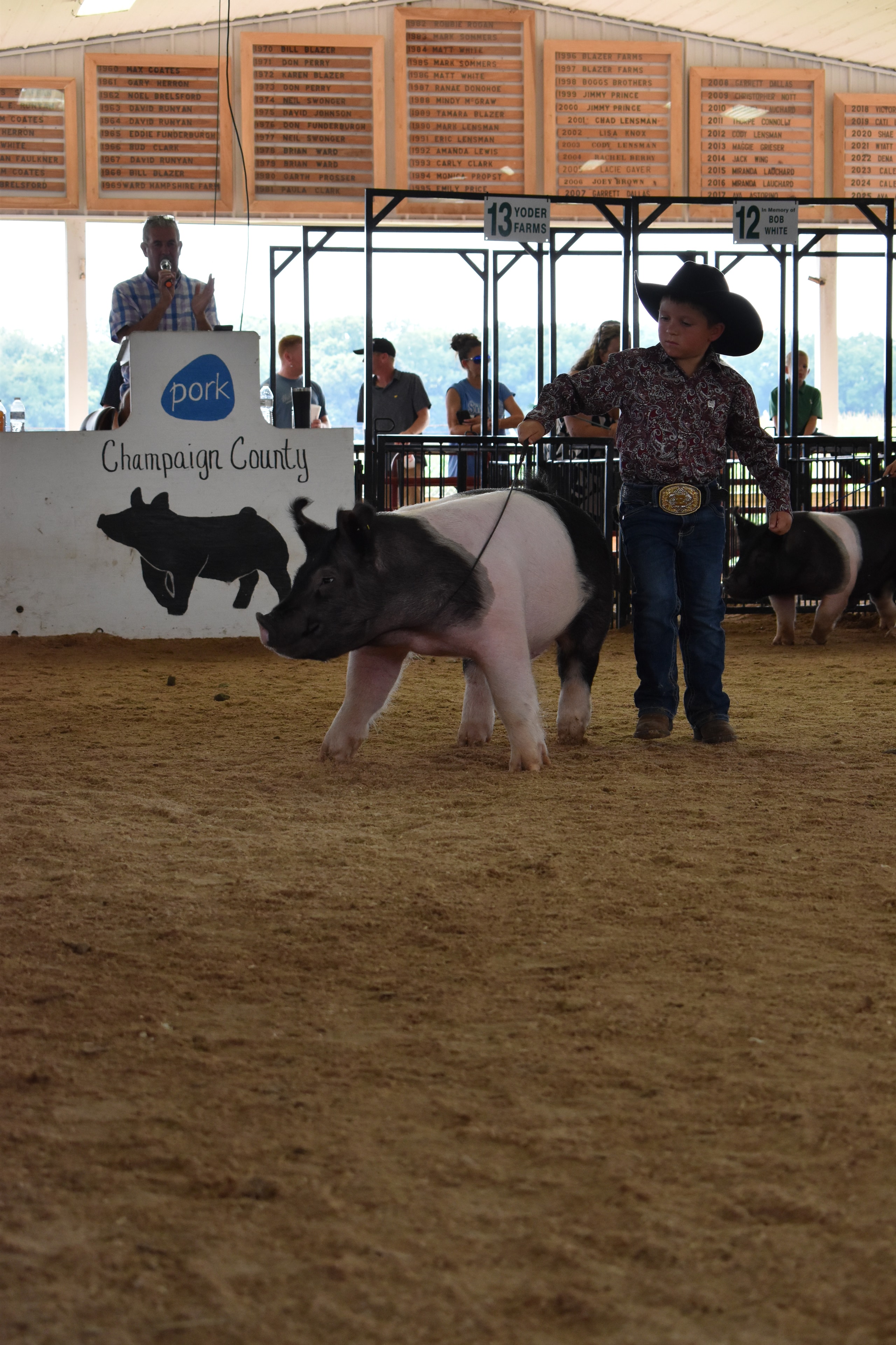 Boy in paisley shirt and black cowboy hat steering Hampshire cross pig with Champaign County Pork sign and announcer at podium