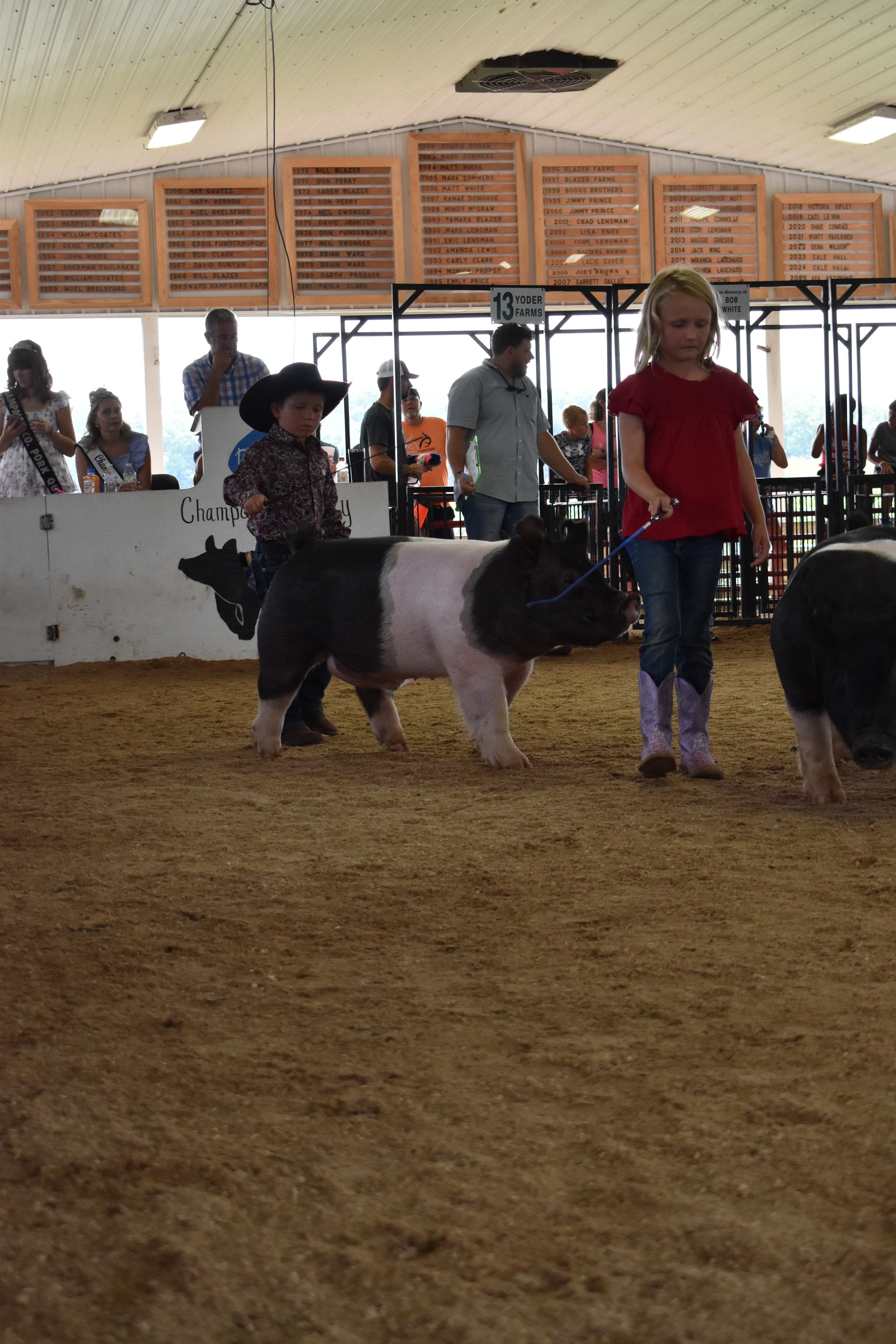 Boy in paisley shirt and black cowboy hat steering Hampshire cross pig past Champaign County Pork sign with Pork Queen and boy in pink polo in background
