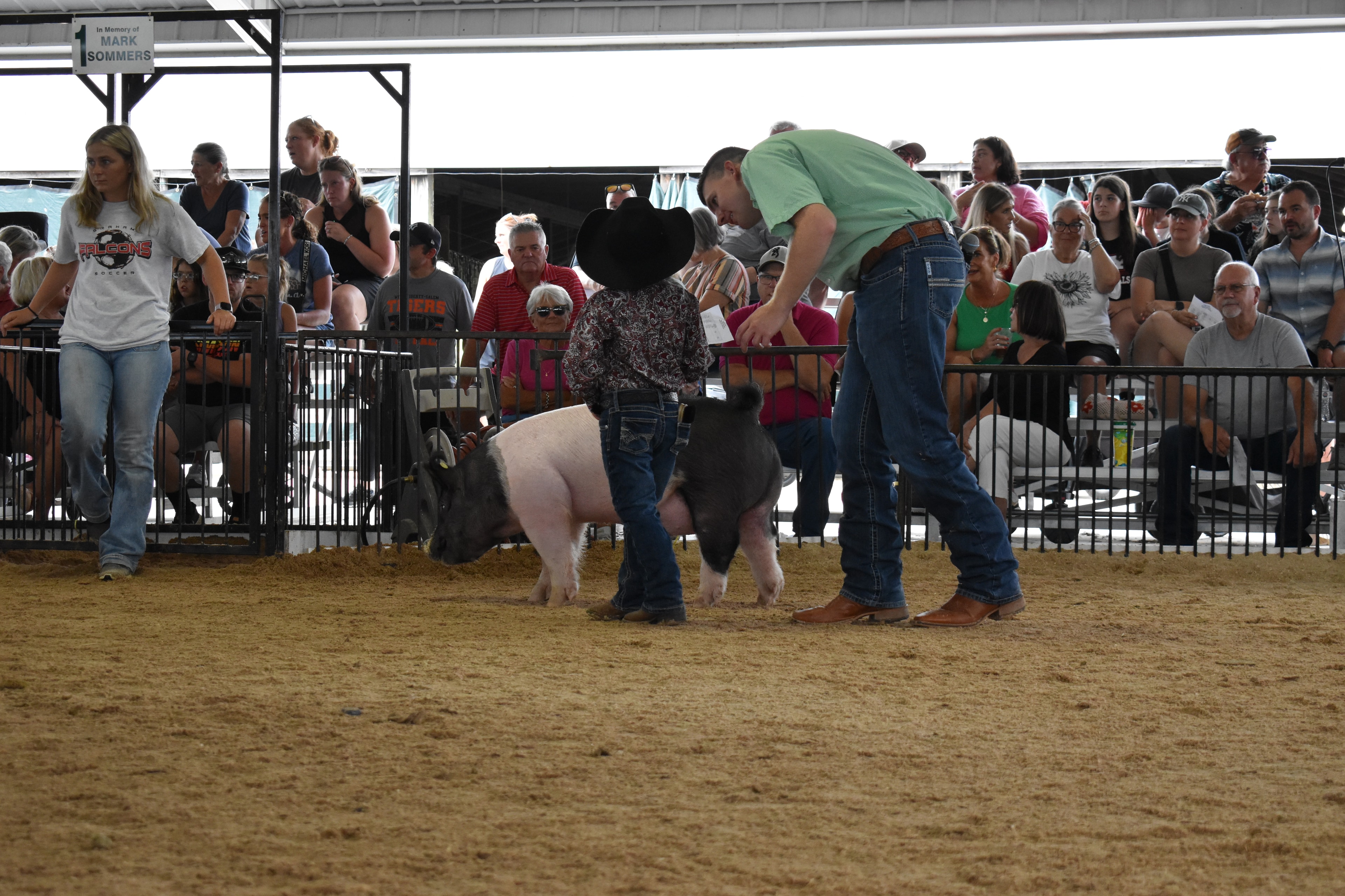 Boy in green shirt steering large black-and-white Hampshire cross pig with girl in yellow shirt and second Hampshire cross pig alongside — Mark IV Farm, J.B.S. United, Da Ja View Farms, Pork Council Women, Crowder Family Farms sponsor pens behind with packed grandstands