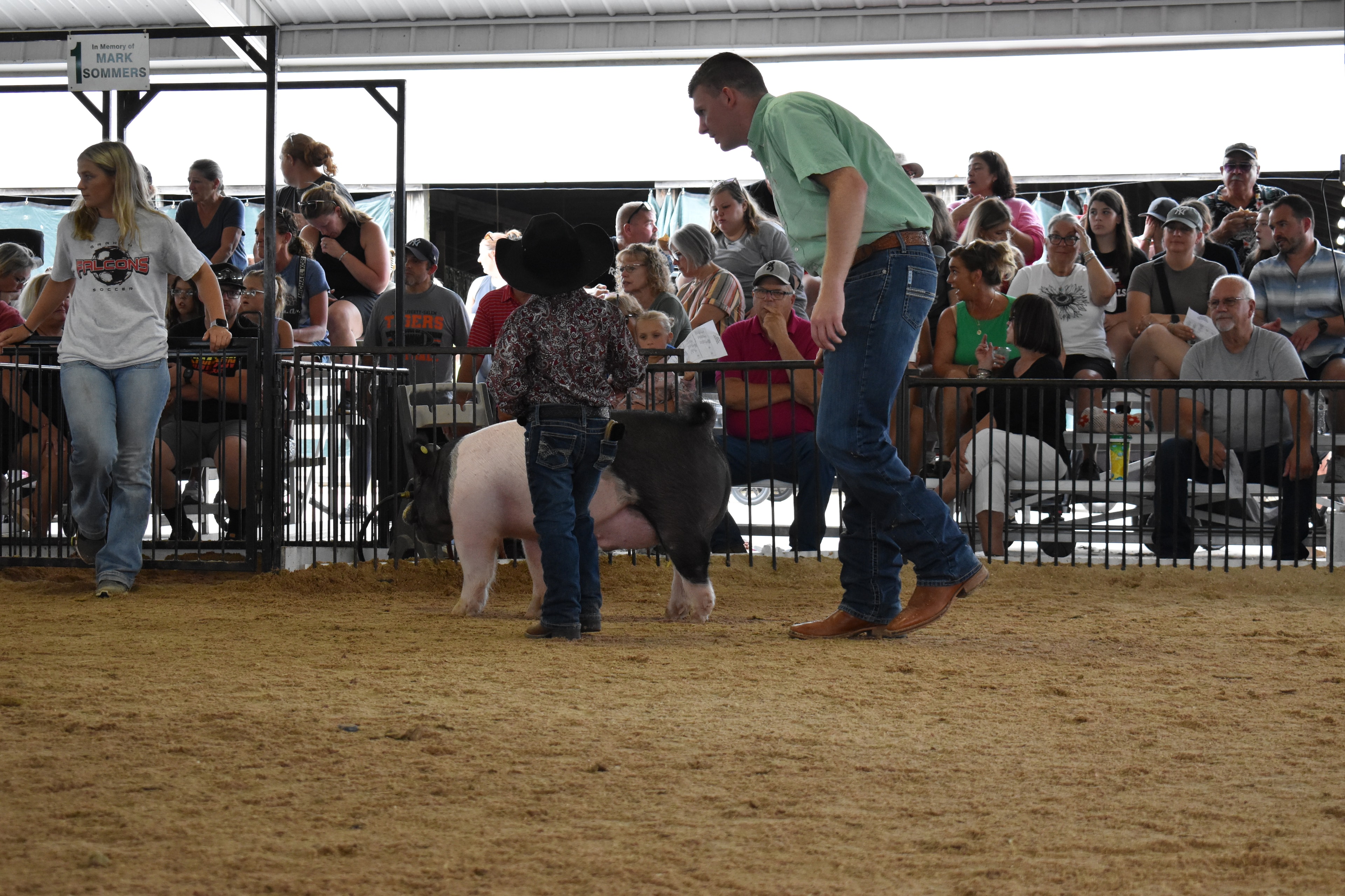 Boy in green shirt steering large black-and-white Hampshire cross pig with girl in yellow shirt and second Hampshire cross pig alongside — Mark IV Farm, J.B.S. United, Da Ja View Farms, Pork Council Women, Crowder Family Farms sponsor pens behind with packed grandstands